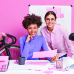 Two diverse creative women collaborate together for making blueprints work on new project pose at coworking space enjoy their occupation isolated over pink background. Women creative workers in office